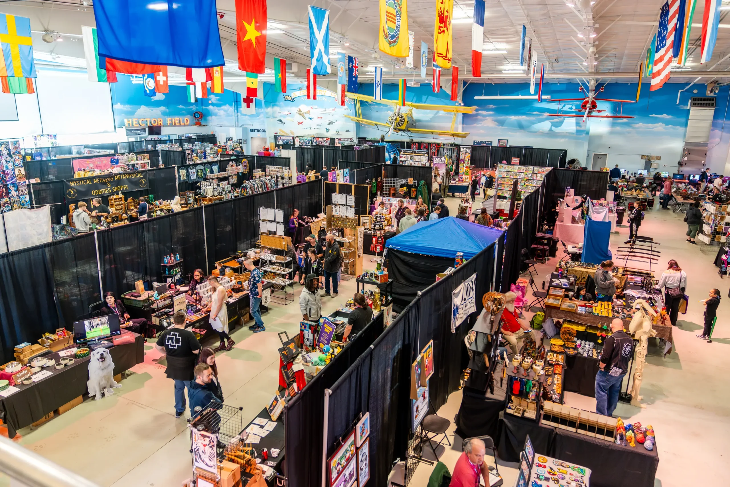 ValleyCon 2025 vendor hall - ValleyCon A busy indoor convention hall with many vendor booths along black curtains and colorful flags overhead. People browse tables of merchandise.